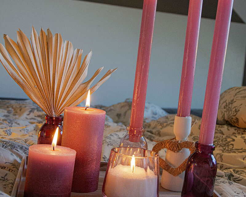 Kerzenarrangement zu Muttertag. Ein weißes Kerzentablett mit Bolsius Rustik Kerzen in verschiedenen Größen, dekorative Stabkerzenhalter aus rosa Glas und weißem Porzellan, mit Rustik-Stabkerzen in der Farbe Eschenrose. Im Vordergrund brennt ein Sandra Rich Teelichtglas, befüllt mit Wenzel Wachsgranulat.