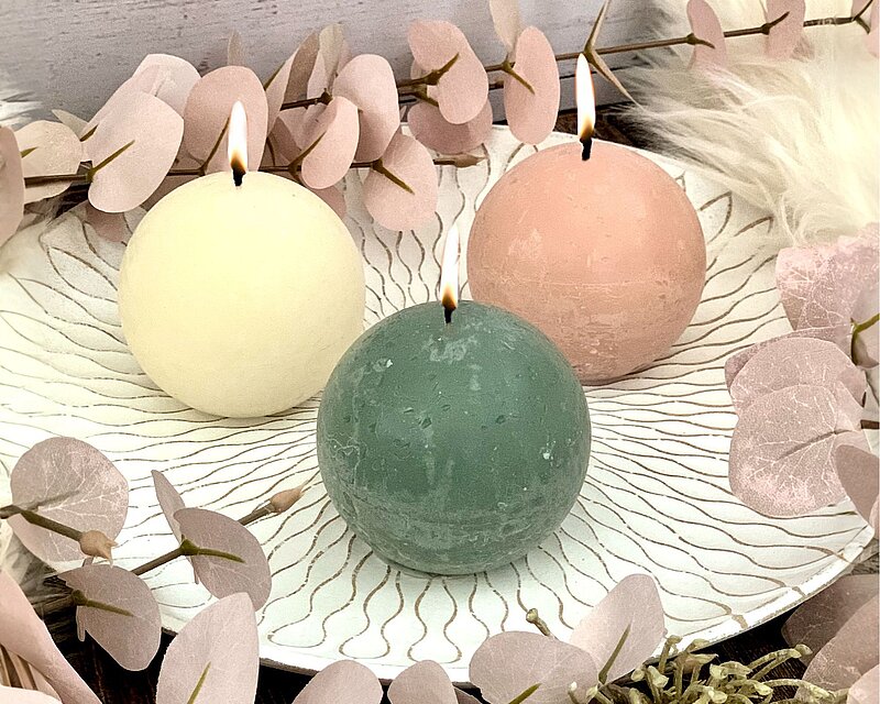 Frühlingsdeko aus einem weißen Holzteller mit goldener Ornament-Verzierung und drei Rustik-Kugelkerzen in den Farben Wolkiges Weiß, Jadegrün und Nebliges Rosa.
