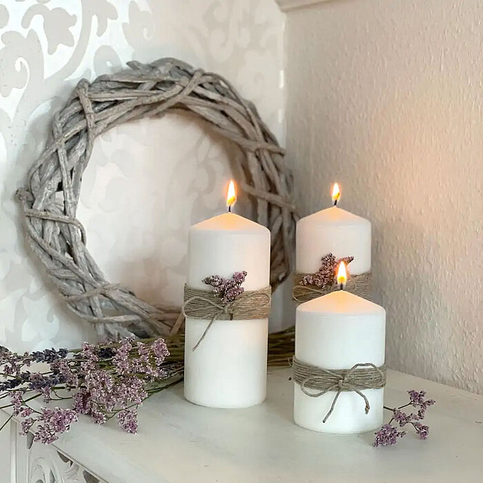Auf einem Holztisch stehen mehrere brennende weiße Teelichter von Müller. Daneben befinden sich zwei brennende weiße Stumpenkerzen von Müller in unterschiedlichen Größen und ein schwarzes, facettiertes Glas, das mit weißen Stabkerzen von Müller gefüllt is