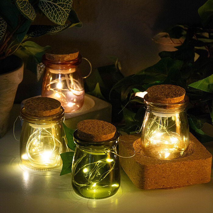 Vier bauchige Glaslaternen vom Hersteller Edelman, beleuchtet durch integrierte Lichterketten, stehen auf einem Holzbrett. Jede Laterne hat einen Korkverschluss und eine Drahtschlaufe zum Aufhängen. Die Laternen sind in den Farben Rosa, Lila, Grün und Kla