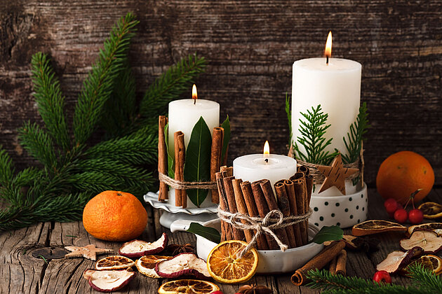 Eine weihanchtliche Szenerie aus weißen Rustik-Kerzen, umgeben von Tannenzweigen, Zimtsternen, Orangen und weihnachtlicher Deko.