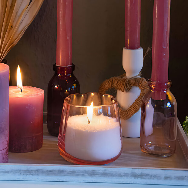 Ein Arrangement aus Kerzen zum Muttertag auf einem weißen Holztablett. Zwei rosa Rustik-Kerzen in verschiedenen Größen, drei Rustik-Tafelkerzen in rosa, die in Kerzenhaltern aus Glas und Keramik stecken. Im Vordergrund ist ein selbstgemachtes Kerzenglas aus Wachsgranulat.