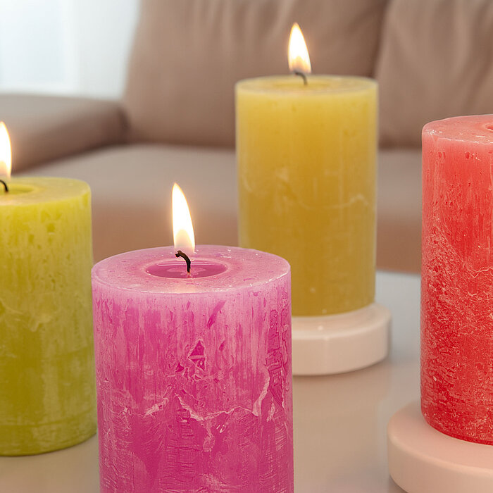 Fünf brennende "DecoLite" Stumpenkerzen in verschiedenen Farben (Weiß, Gelb, Orange und Pink) stehen auf einem weiß gestrichenen Holzregal. Daneben liegen gelbe Blumen als Dekoration. Der Hintergrund ist in warmen, rosafarbenen Tönen gehalten.