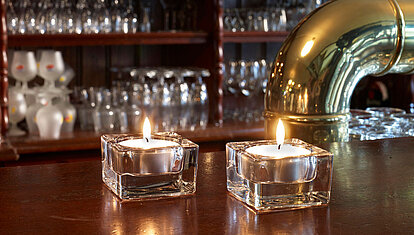 Auf einer Gastronomischen Bar stehen zwei eckige transparente Glas-Teelichthalter mit je einem brennenden weißen Teelicht. Im Hintergrund sieht man mehrere verschiedene Gläser und am rechten Bildrand erkennt man einen goldenen Zapfhahn.