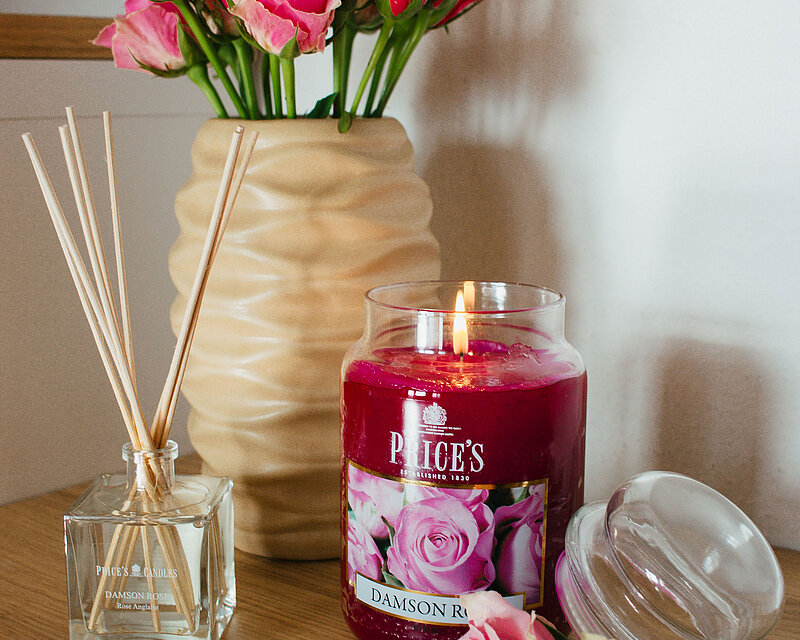 Ein liebevoll gedeckter Tisch zum Muttertag. Im Hintergrund steht eine cremefarbene Keramikvase von Sandra Rich, mit pinken Rosen darin. Im Vordergrund steht ein pinkes Duftglas und ein Raumduft Diffuser von Prices Candles, mit der Duftrichtung "Damson Rose".