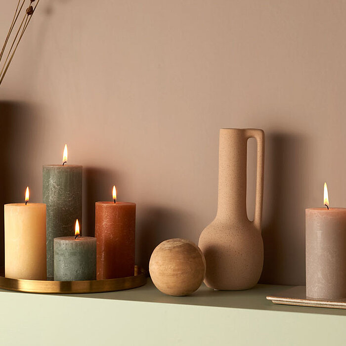 Sechs brennende Bolsius Rustikkerzen in verschiedenen Größen und Farben (Creme, Grün, Rot, Beige, Braun und Grau) stehen auf einer hellgrünen Ablage. Die Kerzen sind teils auf einem goldenen Tablett arrangiert und teils auf der Ablage platziert. Daneben s