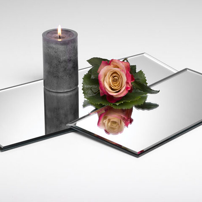 Ein brennendes Teelicht in einem matten, weiß gewellten Glasbehälter steht auf einer quadratischen Spiegelplatte. Daneben befindet sich ein gefrosteter Glasbehälter mit der Aufschrift "Sandra Rich". Um die Kerzen sind zwei silberne, filigrane Dekokugeln p