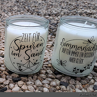 Auf dem Bild stehen zwei Bolsius Starlight Gläser auf einem Kiesboden. Die transparenten Gläser sind mit einem schönen Spruch verziert, der mit Wasserschiebefolie angebracht wurde.