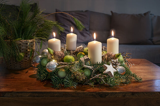  In einem gemütlichen Wohnzimmer steht auf einem Holztisch ein prachtvoller, weihnachtlicher Kranz aus frischen Tannenzweigen, Holzsternen und verschiedenfarbigen grünen und silbernen Kugeln. Auf dem Kranz befinden sich vier brennende, elfenbeinfarbene Stumpenkerzen, die ein warmes Licht verströmen. Im Hintergrund, neben dem Kranz, steht eine schöne Vase aus Bast, gefüllt mit üppigen Fichtenzweigen. Das Bild ist dunkel gehalten und strahlt eine gemütliche Ruhe aus, die an eine friedliche und besinnliche Weihnachtszeit erinnert.