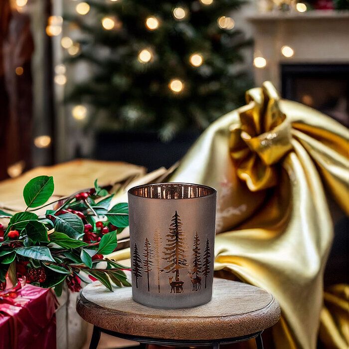 Ein Edelman Teelichthalter mit Baum- und Hirschmotiv steht auf einem kleinen Holzhocker im Vordergrund. Umgeben wird er von weihnachtlicher Dekoration, wie roten Geschenken, grünen Zweigen mit roten Beeren und goldenem Stoff. Im Hintergrund steht ein Weih
