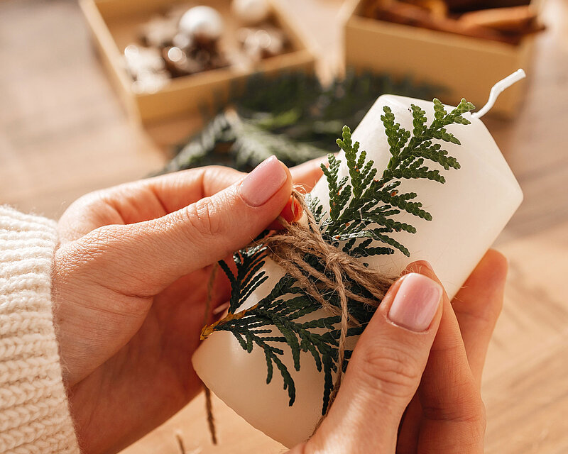 Auf einem Tisch stehen mehrere Bastelmaterialien. Man sieht wie eine Frau eine Stumpenkerze in der Hand hält und mit einer Jutekordel ein Fichtenzweig an der Kerze anbringt. 