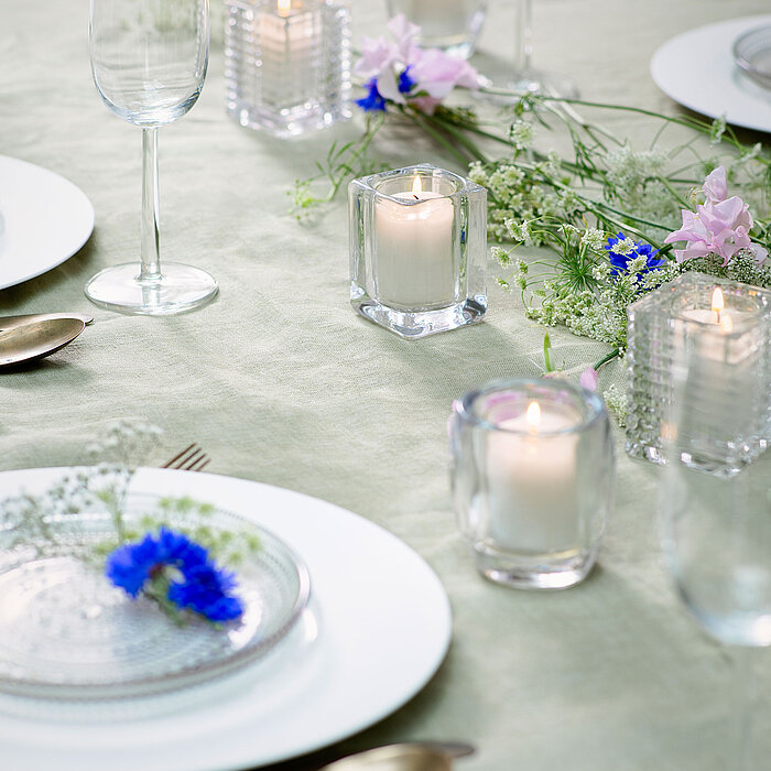 Ein festlich gedeckter Tisch, auf der hellgrünen Tischdecke stehen mehrere brennende Bolsius Clear Cups( Teelicht-Becher) in klaren Glashaltern. Zwischen den Kerzen sind zarte Blumenarrangements in Rosa, Blau und Weiß dekorativ verteilt.