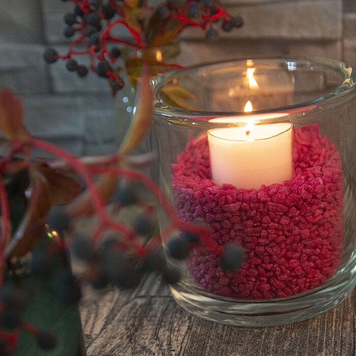 Eine weiße brennende Kerze steht in einem Glasbehälter, der mit pinken Kieselsteinen gefüllt ist. Links daneben befindet sich eine dunkelgrüne Vase mit roten und dunkelblauen Beerenästen. Der Hintergrund besteht aus grauen Steinfliesen, und der Boden hat 