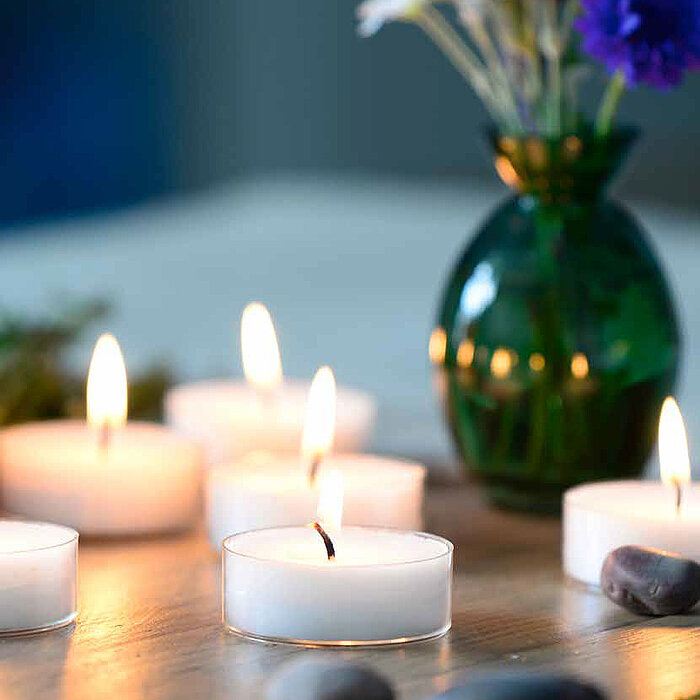 Sechs brennende Teelichter, die auf einem Holztisch arrangiert sind. Daneben liegen einige glatte Steine und eine grüne Vase mit weißen und violetten Blumen. Der Hintergrund ist unscharf, was eine gemütliche und ruhige Atmosphäre vermittelt.