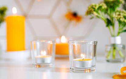 Im Vordergrund steht ein brennendes, gelbes Duft Teelicht in einem Glashalter. Im Hintergrund sieht man weitere gelbe Duftkerzen brennen, sowie eine Vase mit Blumen.