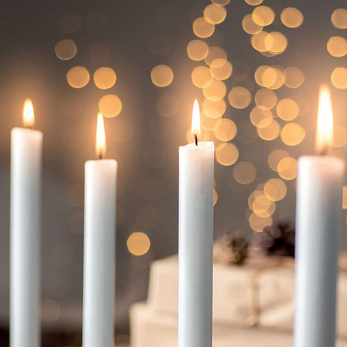 Fünf brennende weiße Stabkerzen vom Hersteller Wenzel stehen in einer geraden Linie. Der Hintergrund ist unscharf und zeigt eine warme, gemütliche Atmosphäre mit goldenen Lichtpunkten und angedeuteten Objekten, die wie Geschenke verpackt sind.