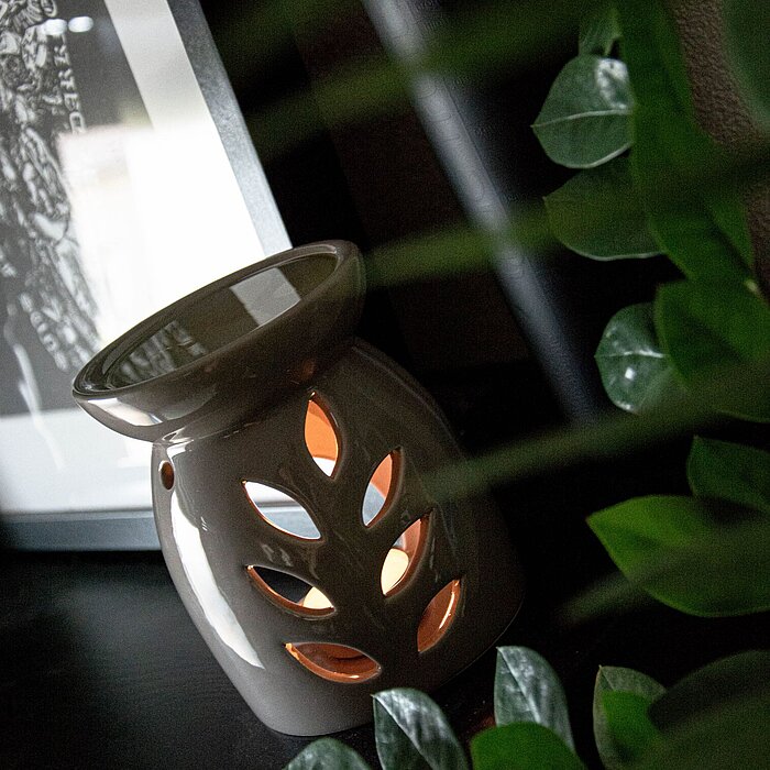 Ein brennender Duftölbrenner in Form einer grauen Vase mit ausgeschnittenen Blattmotiven, durch die das Licht scheint. Der Brenner steht auf einem dunklen Holzregal, neben einer Schwarz-Weiß-Fotografie im Hintergrund. Vor dem Brenner sind grüne Blätter ei