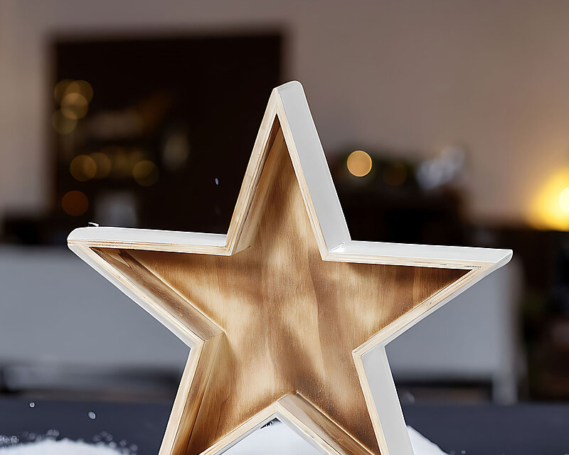 In einem weihnachtlich dekorierten Raum steht auf einer anthrazitfarbenen Kommode ein dekoratives Tablett in Sternform aufrecht. Um den Stern herum ist weißer Kunstschnee verteilt, der eine winterliche Atmosphäre schafft und an verschneite Feiertage erinnert. Die gesamte Szene strahlt festliche Vorfreude und Gemütlichkeit aus.