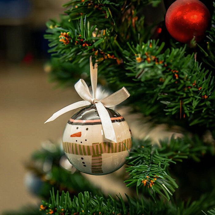 Eine Weihnachtsbaumkugel mit einer Abbildung eines Rentiers hängt an einem grünen Tannenbaum. Der Baum ist mit silbernem Lametta geschmückt, und die Kugel, die in Brauntönen gehalten ist, hat ein stilisiertes Rentiergesicht mit Geweih aufgemalt. Die Kugel