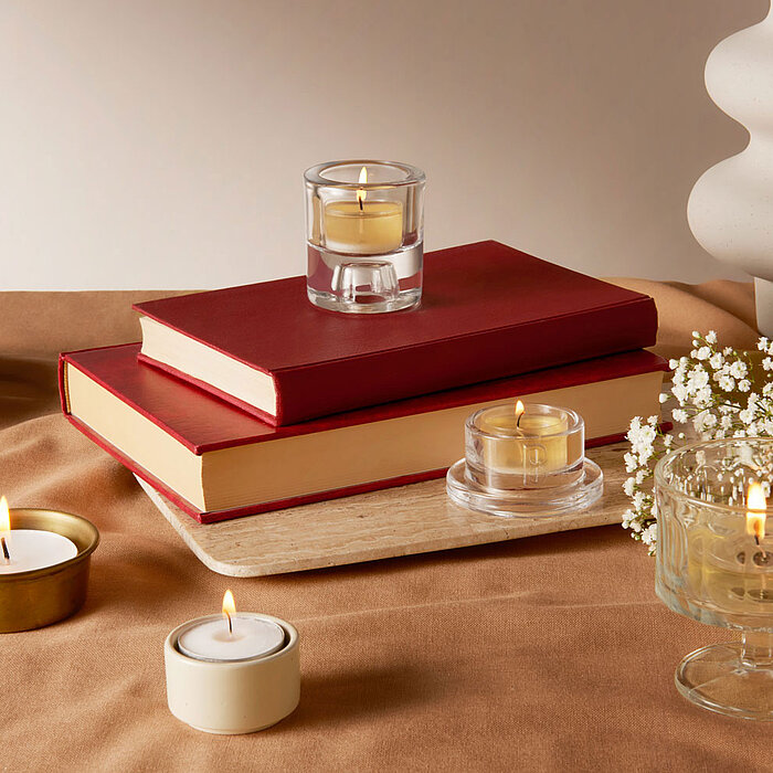 Mehrere brennende Teelichter von Bolsius in durchsichtigen Kerzenhaltern und einem goldenen Becher, arrangiert auf einem beigen, stoffbedeckten Tisch. Im Hintergrund sind zwei rote Bücher zu sehen, eines davon auf einem Holzbrett. Daneben sind weiße Blüte