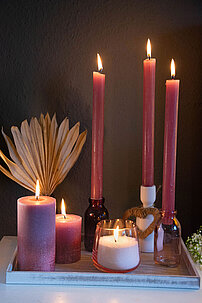 Ein Arrangement aus Kerzen zum Muttertag auf einem weißen Holztablett vor einer grauen Wand. Zwei rosa Rustik-Kerzen in verschiedenen Größen, drei Rustik-Tafelkerzen in rosa, die in Kerzenhaltern aus Glas und Keramik stecken. Im Vordergrund ist ein selbstgemachtes Kerzenglas aus Wachsgranulat.