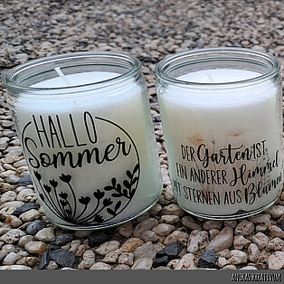 Auf dem Bild stehen zwei Bolsius Starlight Gläser auf einem Kiesboden. Die transparenten Gläser sind mit einem schönen Hallo-Sommer Schriftzug und einem Spruch verziert, die mit Wasserschiebefolie angebracht wurden.