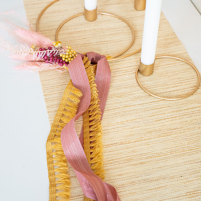 Vier weiße Kerzen in goldenen Kerzenhaltern, die auf einem hellen Tischläufer stehen. Um die Kerzen sind dekorative Bänder in Rosa und Gelb geflochten, verziert mit rosa und gelben Trockenblumen. Die Produkte sind vom Hersteller "Halbach".