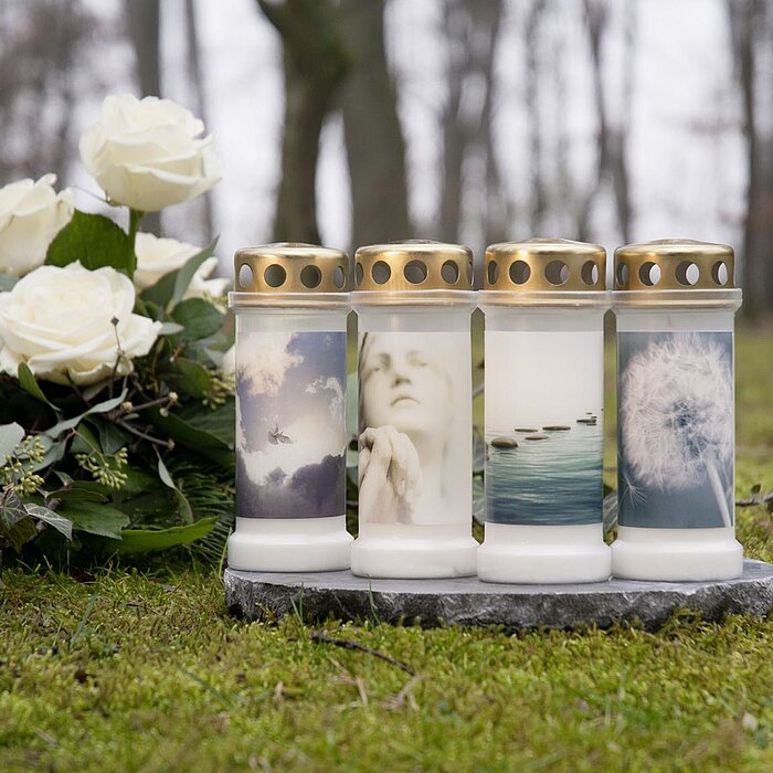Vier Bolsius Grabkerzen mit weißen Wachs in durchsichtigen Glasbehältern stehen nebeneinander auf einer dunklen Steinplatte. Jede Kerze hat einen goldenen Deckel mit Löchern und ist mit verschiedenen beruhigenden Natur- und Seelenbildern bedruckt, darunte