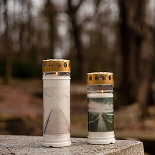 Zu den Grabkerzen Zwei warm leuchtende Grabkerzen mit Motiv und Deckell: Im Fokus steht eine sorgsam platzierte Grabkerze mit goldfarbenem Deckel und stimmungsvollem Himmel‑Motiv. Die Flamme flackert sanft hinter der bedruckten Hülle, umringt von einem Blatt Efeu am Boden des Friedhofs. Hinter dem Motiv‑Grablicht mit Deckel lassen sich weitere Grablichter mit ähnlichem Design erkennen, die im schwindenden Tageslicht leise Gedenken ausdrücken. Die robuste Hülle und der schützende Blechdeckel machen die Grabkerze wetterfest – sie verbindet ein ansprechendes Motiv mit Funktionalität für lange Brenndauer und sicheren Flammenhalt.