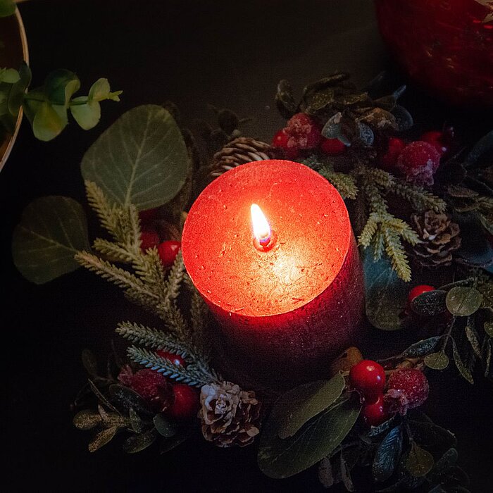 Eine einzelne brennende rote Stumpenkerze steht in einer schwarzen Schale, umgeben von Dekorationsmaterialien wie Tannenzweigen, Beeren und kleinen Zapfen. Die Schale ist auf einem dunklen Tisch platziert, umgeben von grünen Zweigen, die eine gemütliche u