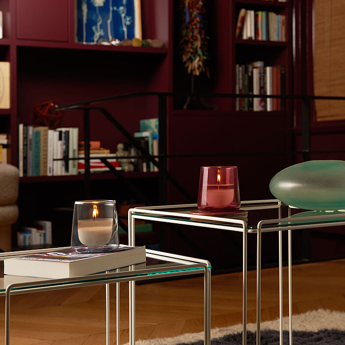 Ein gemütliches Wohnzimmer in warmen Farbtönen. Im Vordergrund stehen zwei Glastische, darauf zwei Bolsius Cleanlight Duftgläser in Grau und Pink.