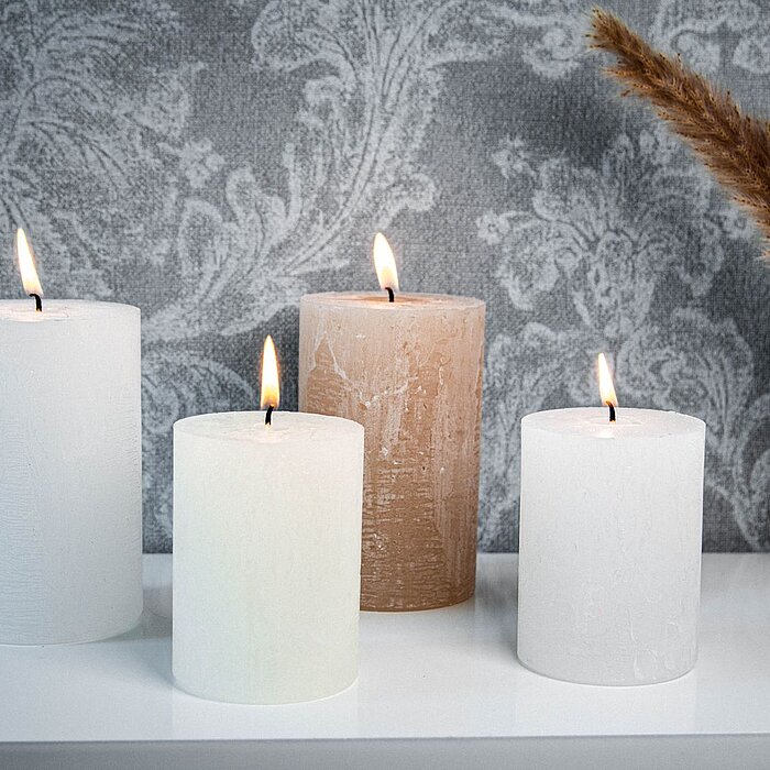 Drei brennende DecoLite-Stumpenkerzen in verschiedenen Höhen stehen auf zwei Holzscheiben, die als Kerzenständer dienen. Die Kerzen haben eine natürliche, beige Farbe und befinden sich auf einer dunklen Holzoberfläche. Im Hintergrund ist eine schlichte Wa