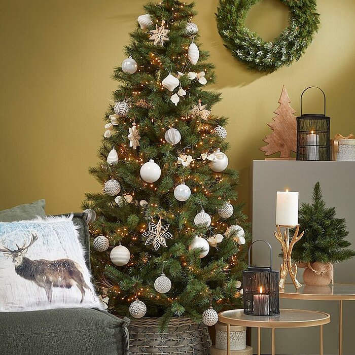 Ein festlich dekorierter Weihnachtsbaum steht in einem modernen Wohnzimmer. Der Baum ist mit weißen Kugeln, goldenen Sternen und Lichterketten geschmückt. Links im Bild befindet sich ein dunkelgrünes Sofa mit einem Kissen, das ein Hirschmotiv zeigt. Recht