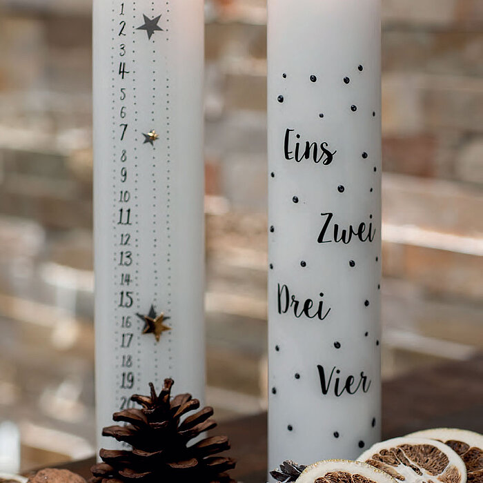 Zwei brennende Stabkerzen des Herstellers Wenzel, die auf einem Tisch dekoriert sind. Eine Kerze ist mit einem Sternenmuster und Zahlen versehen, die als Countdown bis Weihnachten zählen. Die andere Kerze ist mit den Zahlen Eins, Zwei, Drei und Vier sowie