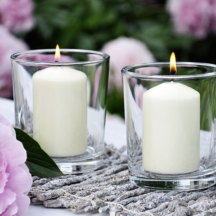 Eine weiße, brennende Stumpenkerze vom Hersteller "Müller" steht auf einem dekorativen, metallischen Tablett mit filigranen Mustern. Daneben befindet sich eine Vase mit einer üppigen, rosa Pfingstrose. Im Hintergrund ist ein grüner, blühender Garten unsch