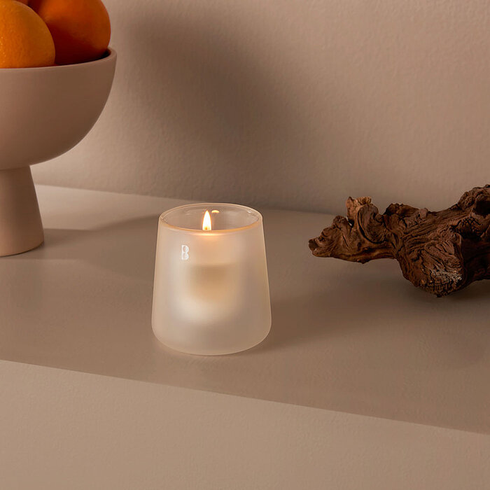 Ein kleiner, brennender Teelicht in einem matten, transparenten Glasbehälter steht auf einer grauen Oberfläche. Daneben liegt ein dekoratives Stück Holz. Im Hintergrund ist eine Schale mit Orangen auf einem Sockel zu sehen. Die Szene wirkt ruhig und minim