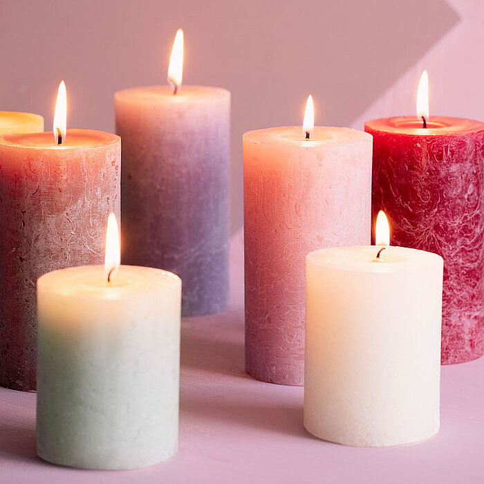 Sieben brennende Bolsius Rustikkerzen in verschiedenen Farben und Größen sind auf einer rosafarbenen Oberfläche angeordnet. Die Farben der Kerzen reichen von Rot, Lila, Rosa, Orange, Weiß bis hin zu Mintgrün, was eine warme und beruhigende Atmosphäre scha