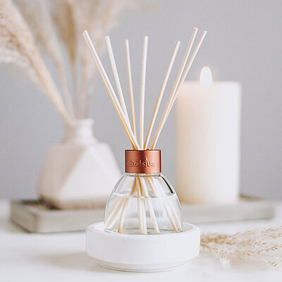Ein freundlicher, heller Raum mit einer hellgrauen Wand. Auf einem weißen Tisch stehen Schilf in einer weißen Vase und eine cremefarbene Stumpenkerze im Hintergrund. Im Vordergrund ist ein eleganter Raumduft Diffuser mit Rattanstäbchen zu sehen. Dieser läßt sich als Alternative zu Duftkerzen einsetzen.