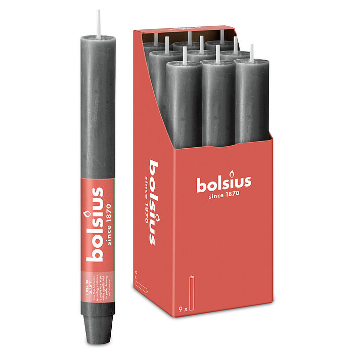 Eine durchgefärbte XL Rustik Haushaltskerze mit konischem Fuß in der Farbe Stürmisches Grau. Im Hintergrund ist ein 9er Pack der Stabkerzen in einer Schachtel von Bolsius zu sehen.