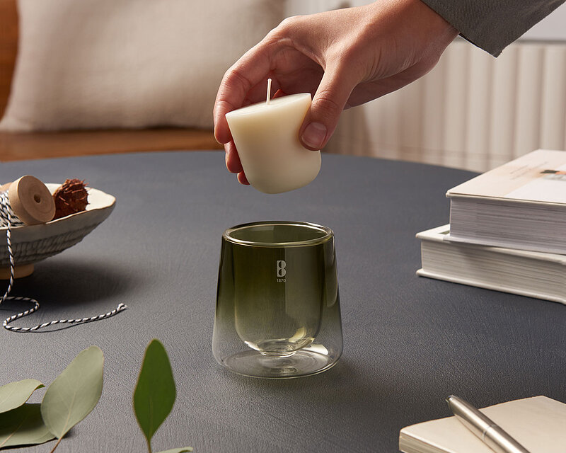 Es ist ein ruhiges und klares Bild mit aufeinander abgestimmten Farbtönen. In einem gemütlichen Wohnzimmer auf einem dunkelgrauen Wohnzimmertisch steht ein Bolsius Cleanlight Glas. Man sieht wie ein Mensch das Cleanlight Nachfüllwachs in dem Glas platzieren möchte. Auf dem Tisch befinden sich mehrere Bücher und ein paar grüne Zweige.  