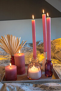 Ein Arrangement aus Kerzen zum Muttertag auf einem weißen Holztablett. Zwei rosa Rustik-Kerzen in verschiedenen Größen, drei Rustik-Tafelkerzen in rosa, die in Kerzenhaltern aus Glas und Keramik stecken. Im Vordergrund ist ein selbstgemachtes Kerzenglas aus Wachsgranulat.