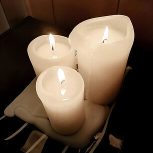 Beispiel für die Gefahren, wenn man brennende Kerzen zu nah nebeneinander aufstellt. Die drei brennenden weißen Stumpenkerzen sind ausgelaufen, das Wachs fließt auf den Kerzenteller darunter.