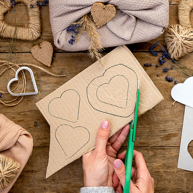 Eine Nahaufnahme ienr Hand, die mit einer Schere Herzen aus Pappe ausschneidet. Im Hintergrund sieht man bereits fertige Deko-Herzen aus Jute zum Muttertag.