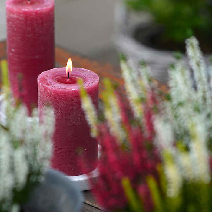 Vier brennende Bolsius Rustikkerzen in den Farben Gelb, Weiß, Grün und Pink stehen auf einem weißen, hölzernen Tisch. Im Hintergrund ist ein rosenholzfarbener Farbton an den Wänden und gelbe Blumen zu sehen, die eine fröhliche Atmosphäre schaffen. Eine we