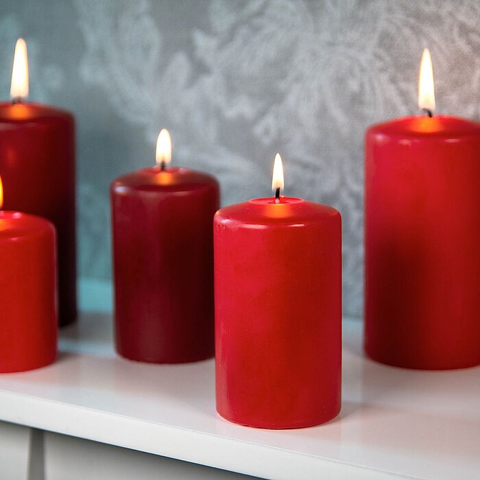 Fünf brennende rote Stumpenkerzen von DecoLite in verschiedenen Größen stehen auf einem weißen Regal. Der Hintergrund zeigt eine graue, floral gemusterte Tapete, die eine elegante Atmosphäre schafft.