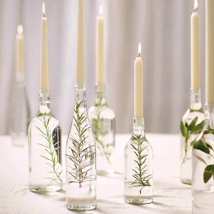 Elf brennende Bolsius Tafelkerzen in elegant arrangierten Glasflaschen auf einem festlich gedeckten Tisch. Die Tischdekoration umfasst grüne Blätter, weiße und rosa Blumen sowie zarte Glaswaren, die in einem filigranen Zusammenspiel eine romantische Atmos