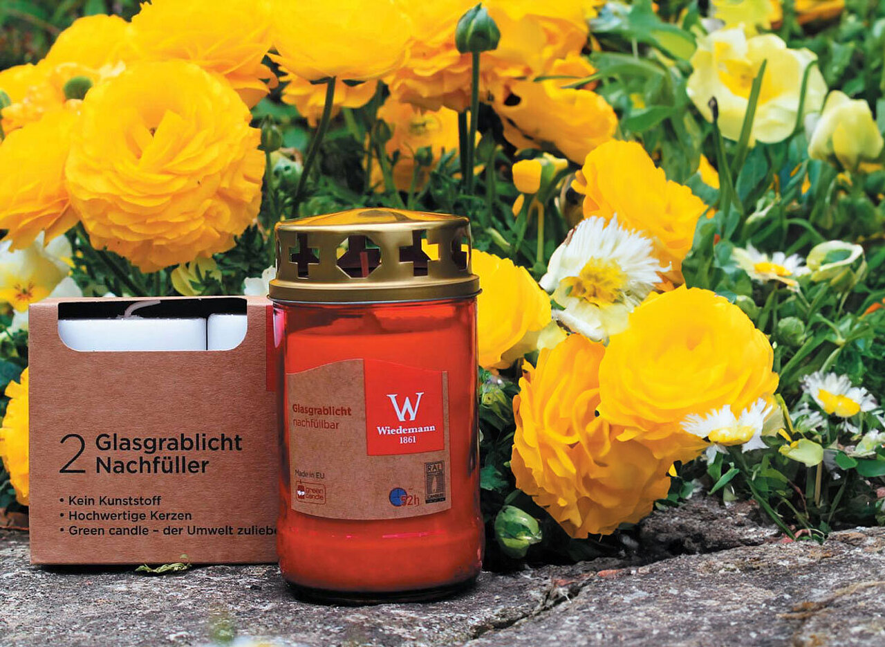 Im Vordergrund auf der linken Seite steht ein Glas-Grablicht aus rotem Glas von Wiedemann. links daneben steht eine braune Verpackung mit zwei weissen Glasgrablicht Nachfüllern von Wiedemann. Dahinter sieht man einige gelbe Ranunkeln und grüne Knospen und einige Gänseblümchen. Alles steht und liegt auf einem Steinboden.