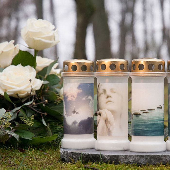 Bei dem Bild sind vier brennende Bolsius Grabkerzen zu sehen. Zwei Kerzen tragen jeweils ein Bild der Jungfrau Maria. Die Kerzen sind auf einem verwitterten Stein platziert, umgeben von herbstlichem Laub. Neben den Grabkerzen steht eine Metalllaterne mit 