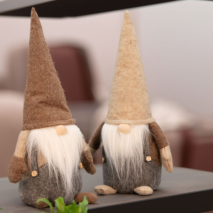 Zwei dekorative Weihnachtswichtel vom Hersteller Edelman stehen auf einem Regal. Sie haben lange weiße Bärte und spitze Hüte, einer in Braun und der andere in Beige. Ihre Körper sind aus grauem Filz gefertigt und sie tragen kleine Knöpfe. Im Vordergrund i