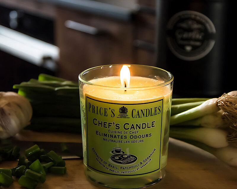 Ein grünes Duftglas Chefs Candle von Prices steht auf einer Holzarbeitsplatte in einer dunkelbraunen Küche. Auf der Holzarbeitsplatte befinden t sich ein Bund Frühlingszwiebeln und ein paar geschnittene Frühlingszwiebeln und ein Knoblauch.